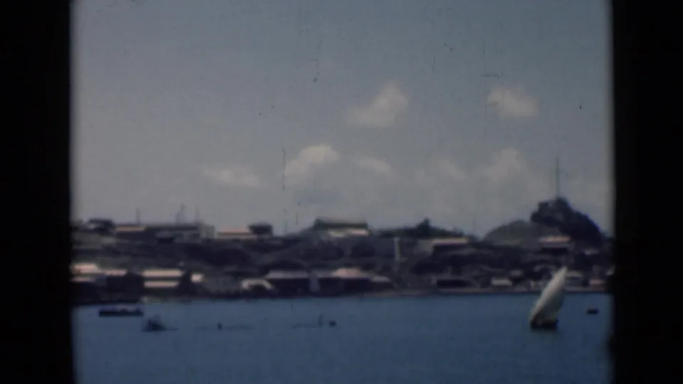 1946 8mm view across Aden harbor with small boats, waterfront buildings, hillside structures, mountains, and clouds.