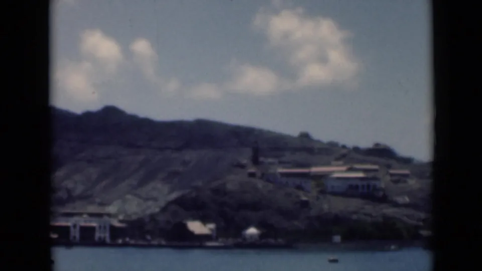 1946 8mm frame showing Aden harbor water, a rocky hillside, scattered waterfront buildings, and partly cloudy sky.