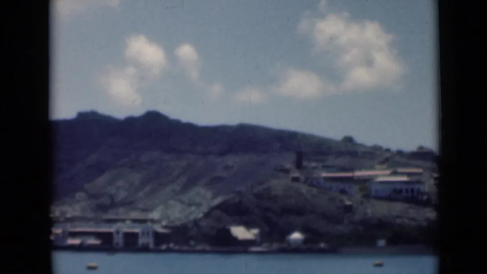 Aden harbor scene on 8mm film, with low shoreline buildings, steep mountain terrain, calm water, and a small boat near the foreground.