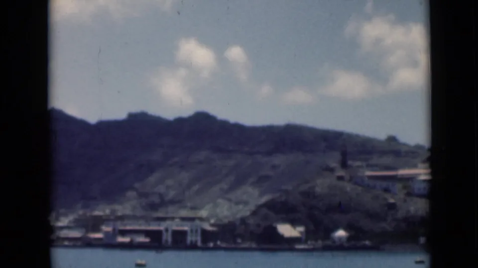 1946 home-movie frame of Aden harbor, showing small boats on blue water, low waterfront structures, dark mountains, and scattered clouds.
