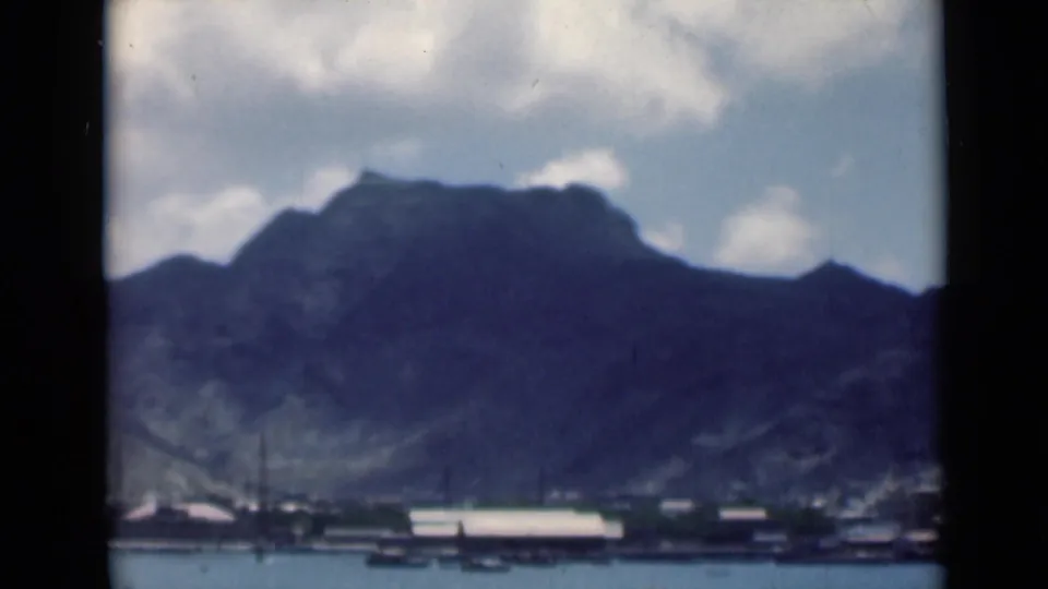 1940s 8mm travel frame showing a shaded mountain slope above a waterfront strip with boats and pale-roofed buildings.