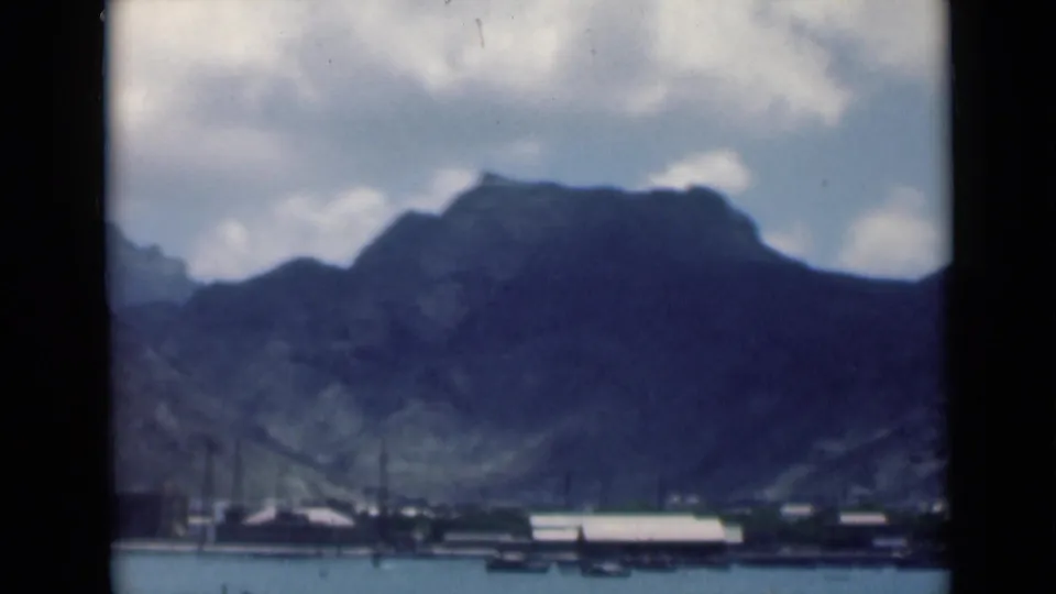 Frame from 1946 8mm footage identified as Aden, Yemen, with mountains, a cloudy sky, harbor boats, and waterfront structures.