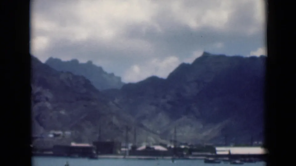 The 8mm frame shows a distant Aden waterfront with calm water, small boats, low structures, and steep shadowed mountains behind.