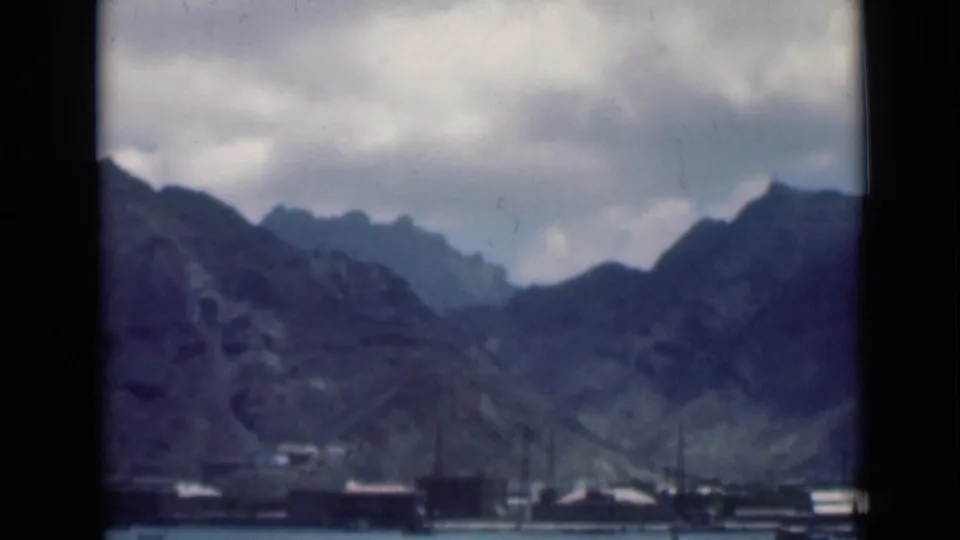 A wide home-movie view of Aden harbor shows low waterfront buildings and moored boats beneath rugged mountains and a cloudy sky.