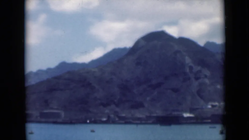 Archival 8mm frame showing a steep mountain slope above blue water, with low structures and small boats near the shore.