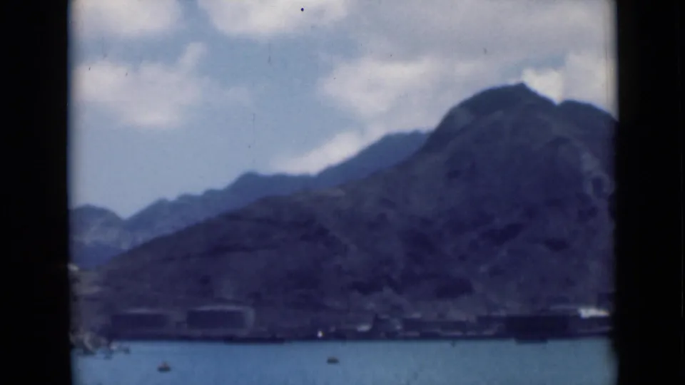 Final 8mm frame of the coastal mountain view, showing blue water, tiny boats, and a rugged shoreline in soft focus.