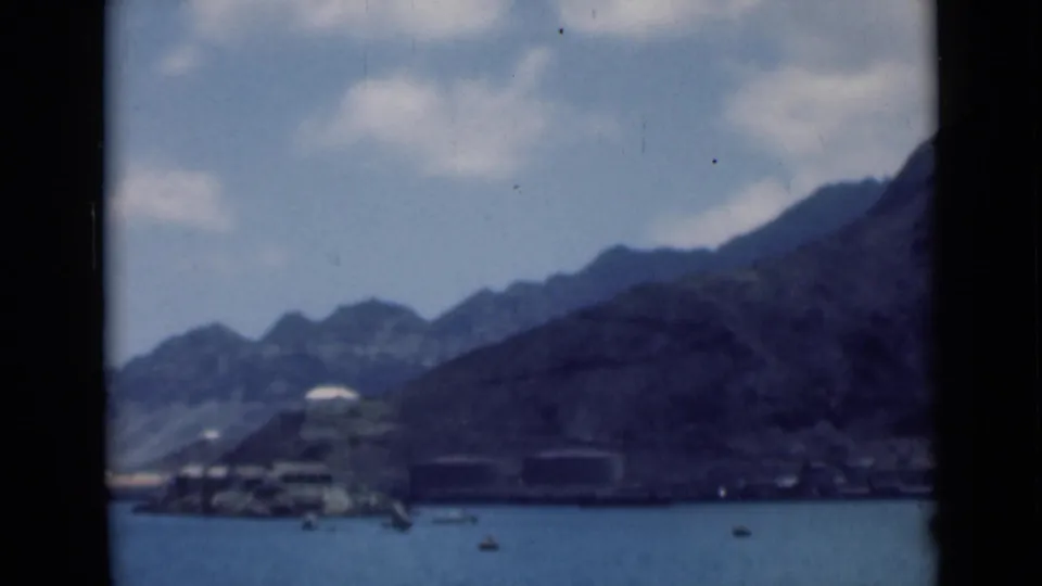 Distant harbor-like shoreline identified by supplied metadata as Aden, Yemen, with boats on calm water and mountains in the background.