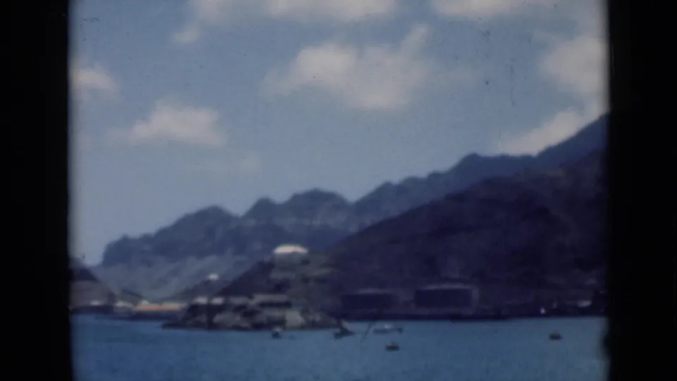 Soft 8mm view of a coastal scene identified as Aden, Yemen, with boats, a rocky shore, low structures, and steep mountains.