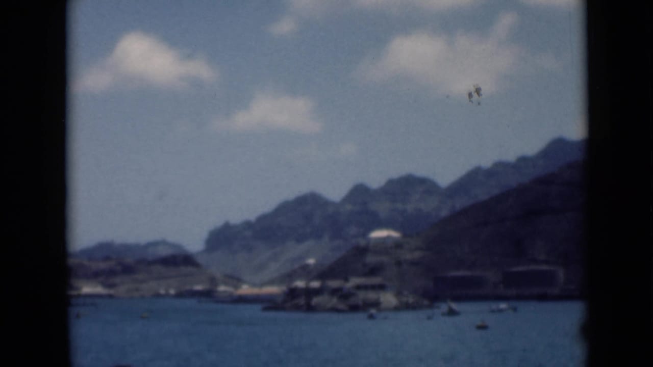 1940s 8mm view consistent with Aden, Yemen metadata, showing calm harbor water, small boats, shoreline structures, and rocky hills.