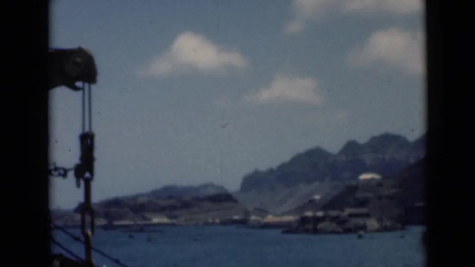 Home-movie harbor view with water, boats, shoreline buildings, rugged hills, and a dark obstruction along the left edge of the frame.