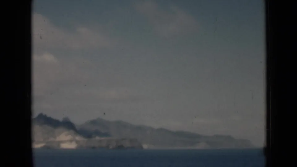 1940s 8mm view of a distant rocky island or headland across calm water near Aden, Yemen, with hazy hills and a cloudy sky.
