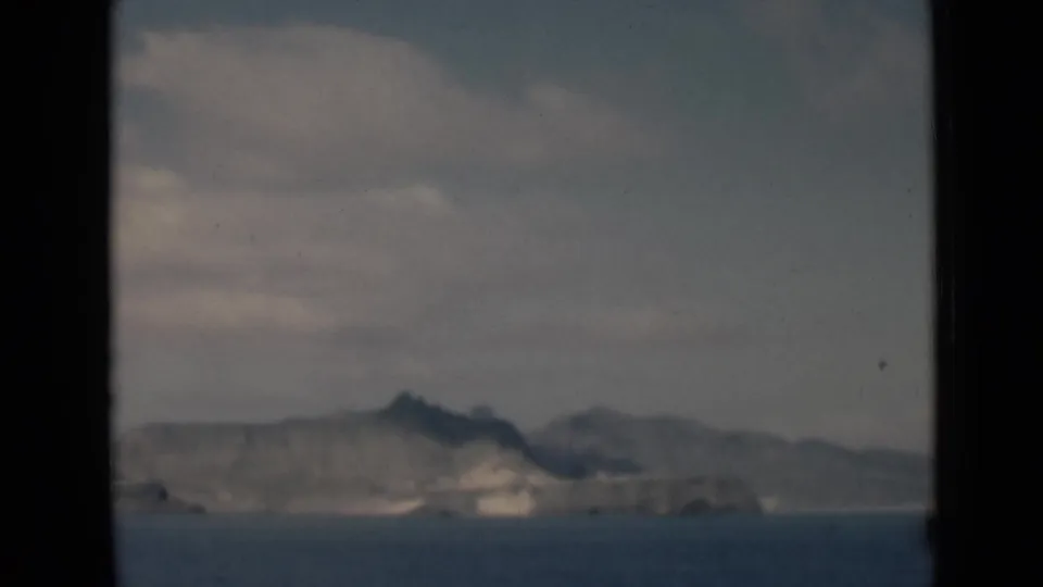 1940s 8mm frame of distant hills beyond blue water, seen through dark window-like borders under pale clouds.