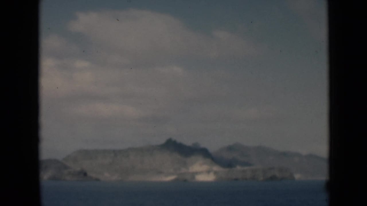 Rocky shoreline and hills across water beneath soft clouds, with a dark vertical frame edge.