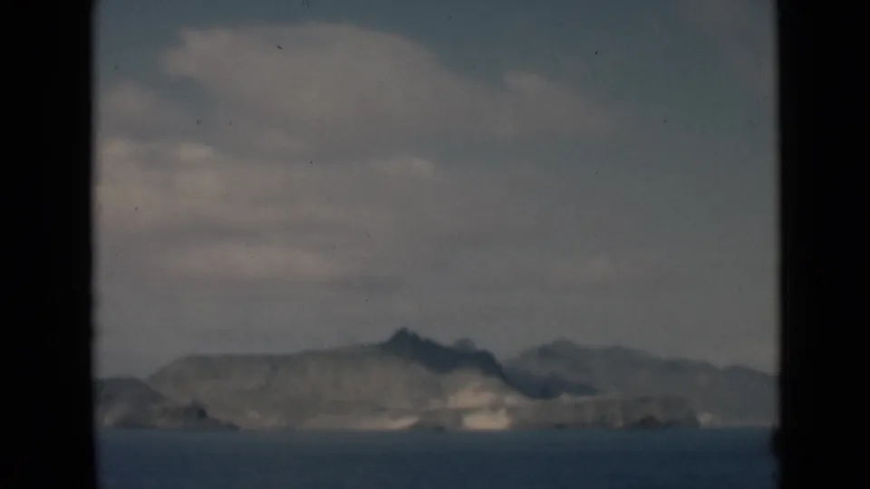 Soft 8mm view of a distant rocky coast or lakeshore across water, framed by a dark vertical edge and cloudy sky.