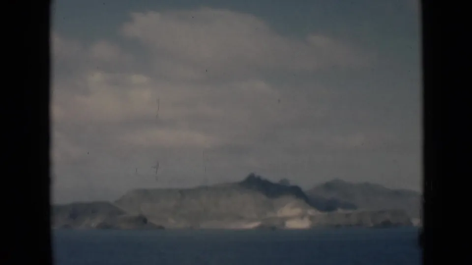 Vintage 8mm frame showing low rocky hills and a pale shoreline across blue water below a clouded sky.