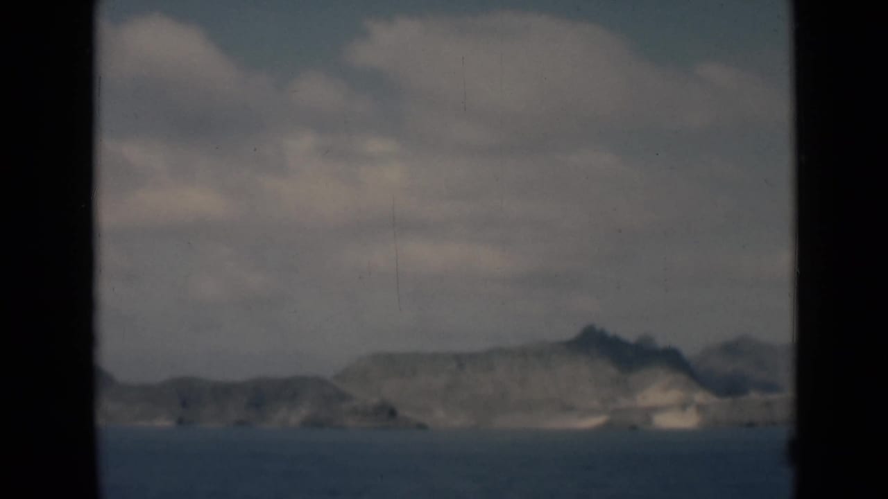 Distant rocky coastline across calm water under a cloudy sky, framed by dark film borders.