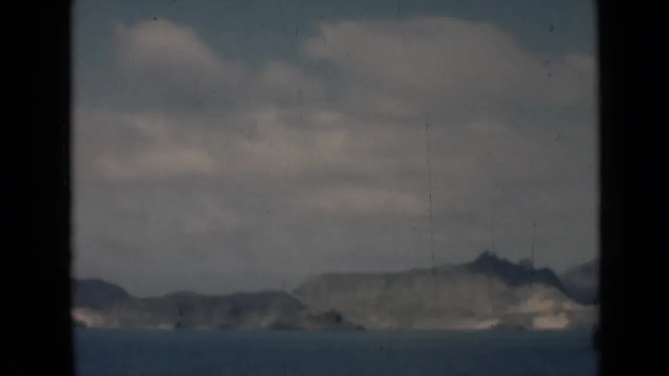 A soft 8mm view of the Aden coastline, showing rocky cliffs beyond calm water beneath a pale, clouded sky.