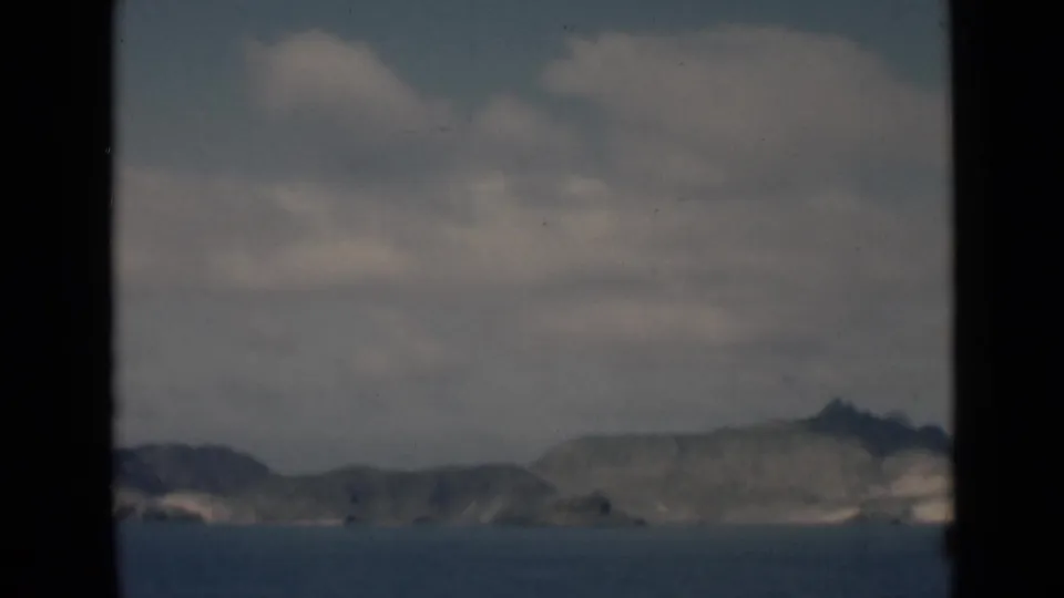1946 home-movie frame of a low rocky shoreline near Aden, with open water in the foreground and clouds overhead.
