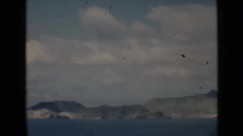 A distant Aden coastal view on 8mm film, with calm water, rocky shoreline, clouds, and small dark birds or film marks in the sky.