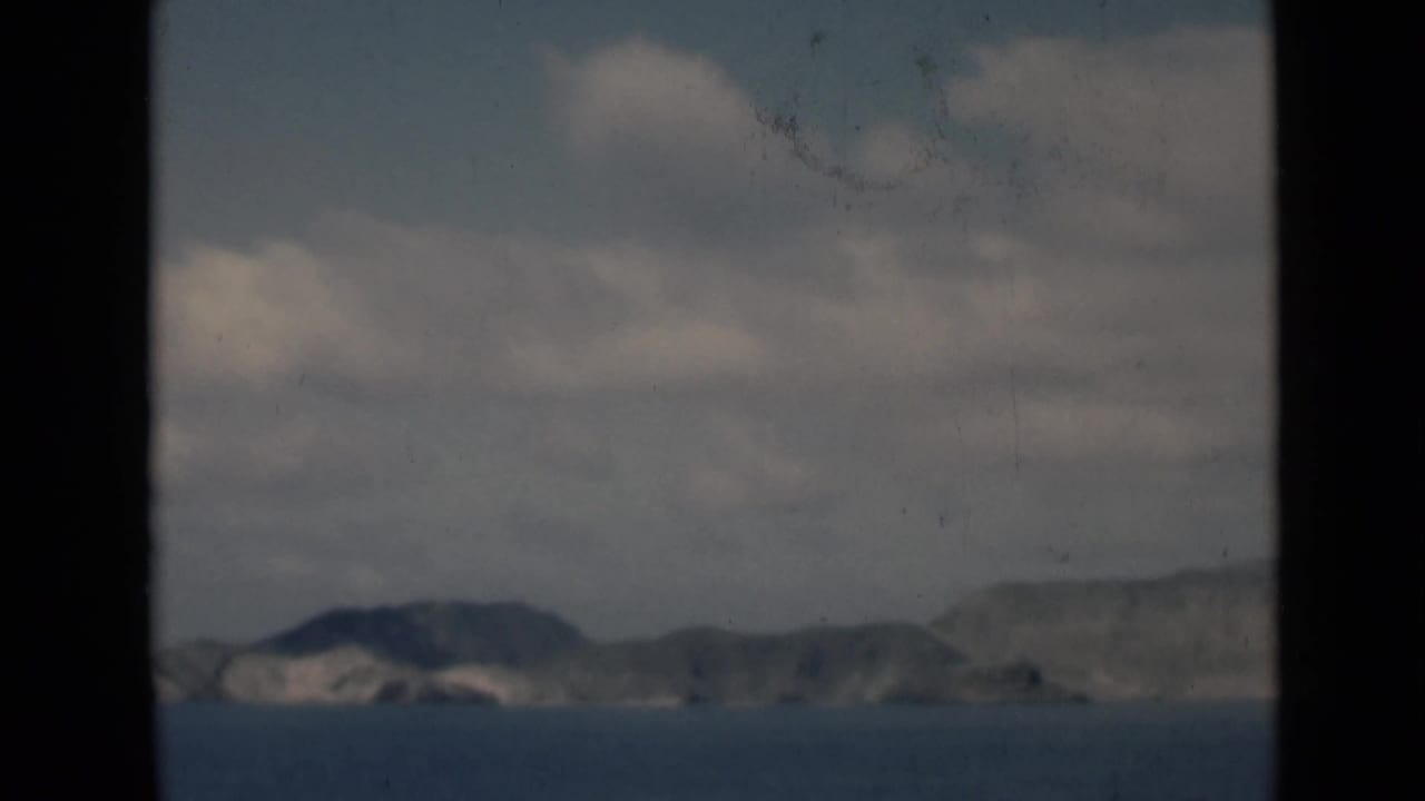 Distant coastal mountains across blue water beneath a cloudy sky, framed by dark vertical borders.
