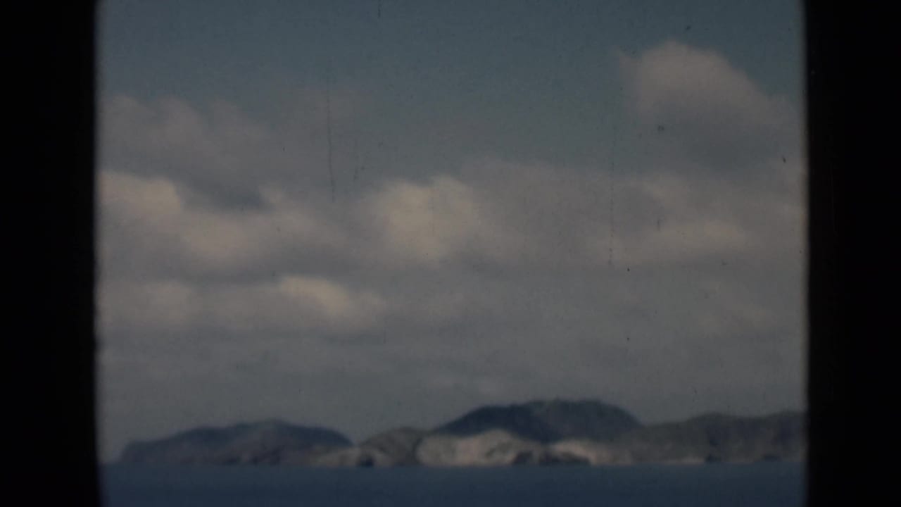 Distant rocky shoreline across blue water under cloudy sky, seen through dark window framing.