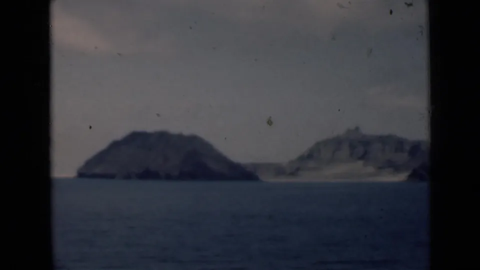 1946 home-movie frame showing a small dark island offshore with pale rocky coastal land beyond it.