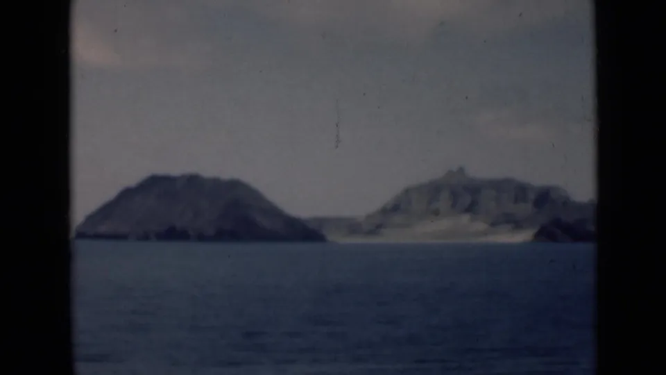 A 1940s 8mm shipboard view shows two rocky islands across calm blue water, with dark film borders and a hazy sky.