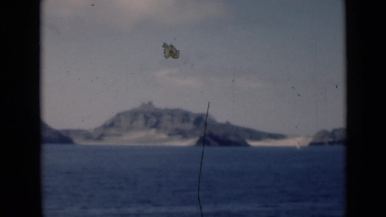 1946 8mm frame showing a distant rugged island and pale shoreline seen from across open water near Aden, Yemen, per supplied metadata.