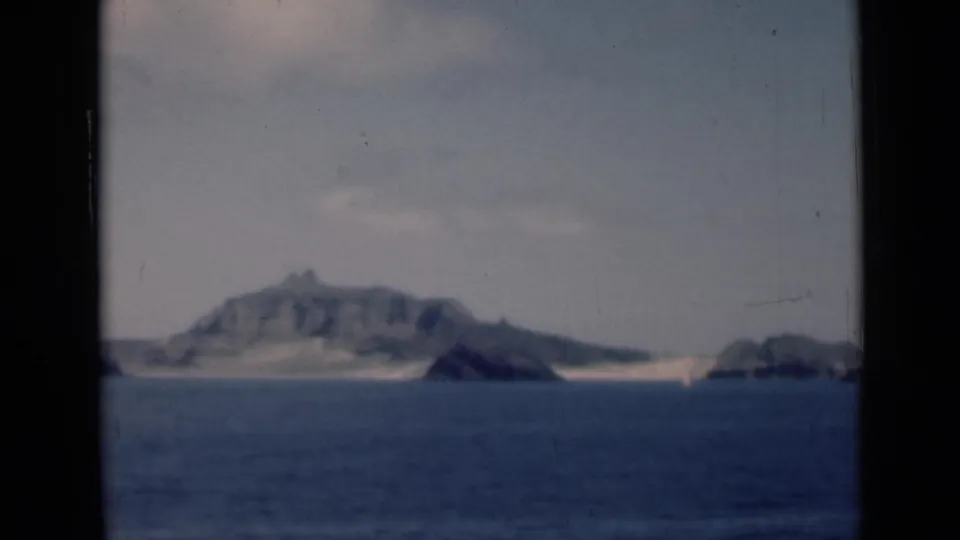 A shipboard 8mm view of a rugged island and low shoreline across blue water, with dark gate edges and muted color.