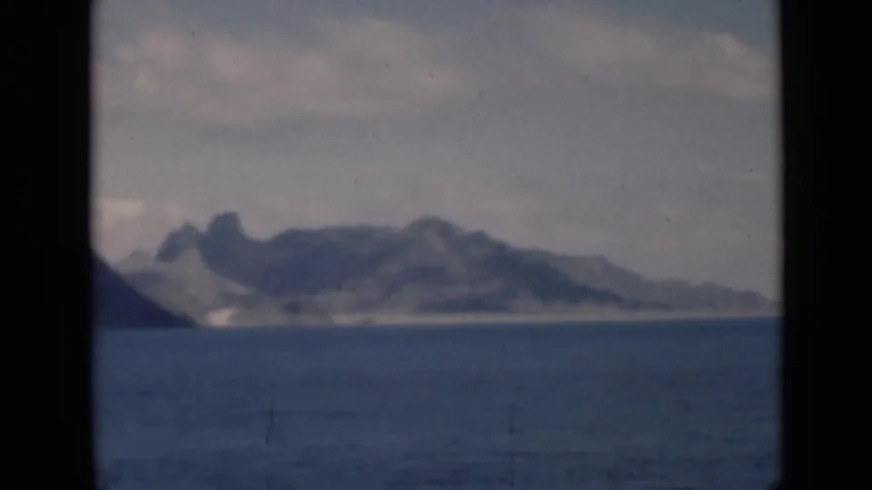 1946 8mm frame of a hazy mountainous coastline across calm water, with dark frame borders and soft focus.