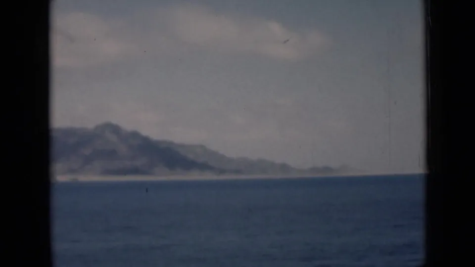 Soft 8mm view of a low mountain coastline and pale shore across rippling water during a boat passage.