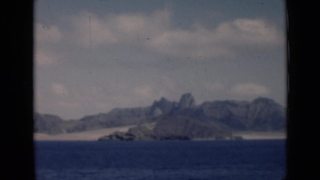 1946 8mm home-movie view toward a distant Aden/Yemen-associated coast with blue sea, pale dunes, hazy sky, and rugged mountains.
