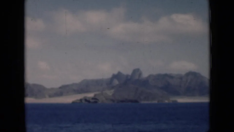 1946 8mm home-movie view toward a distant Aden/Yemen-associated coast with blue sea, pale dunes, hazy sky, and rugged mountains.