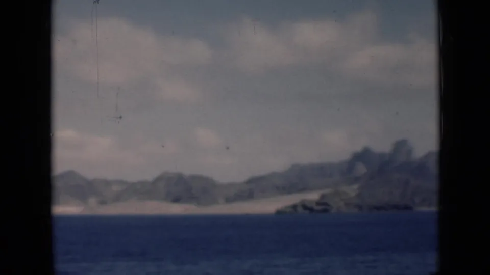 The camera holds on a distant coastal landscape associated with Aden/Yemen, showing sea, sandy shore, mountains, haze, and heavy film softness.
