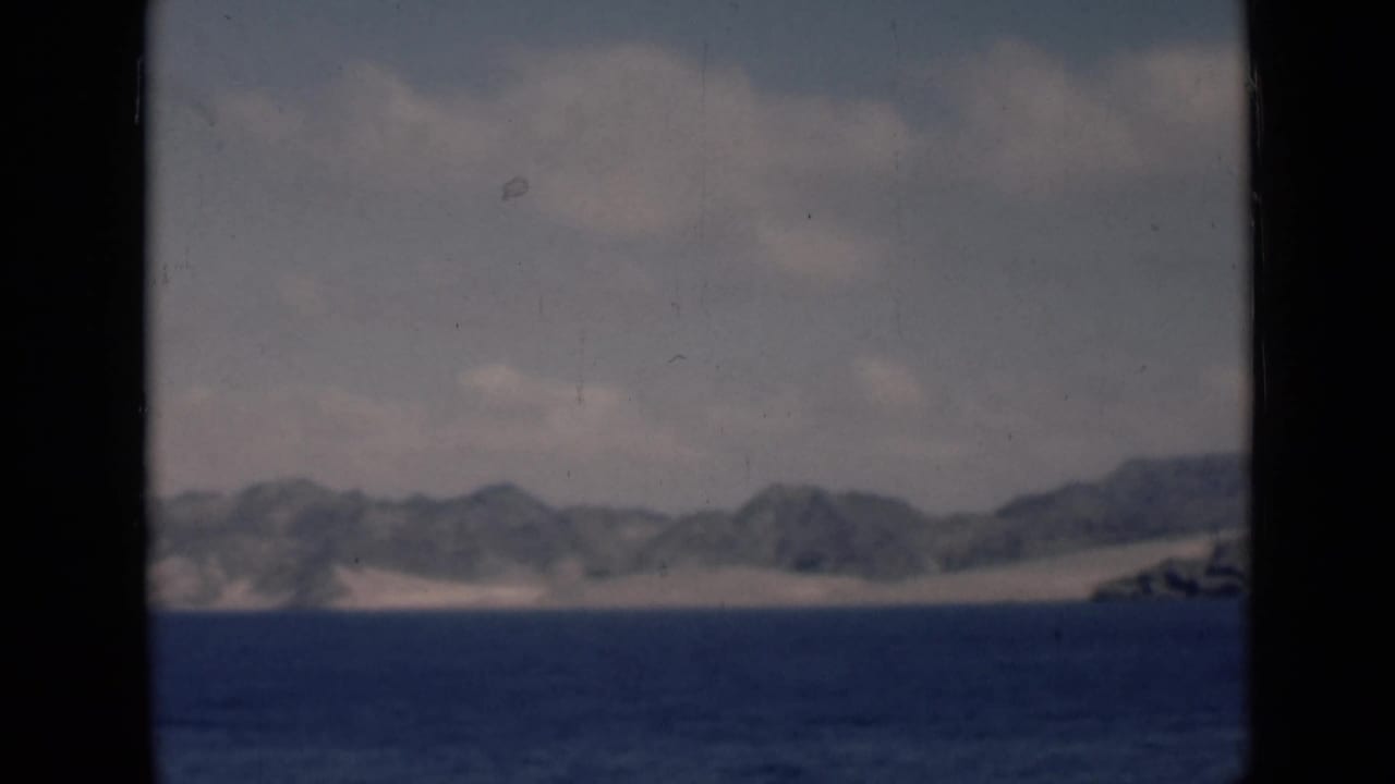 Distant arid mountains and sandy shore across blue water under a pale cloudy sky.