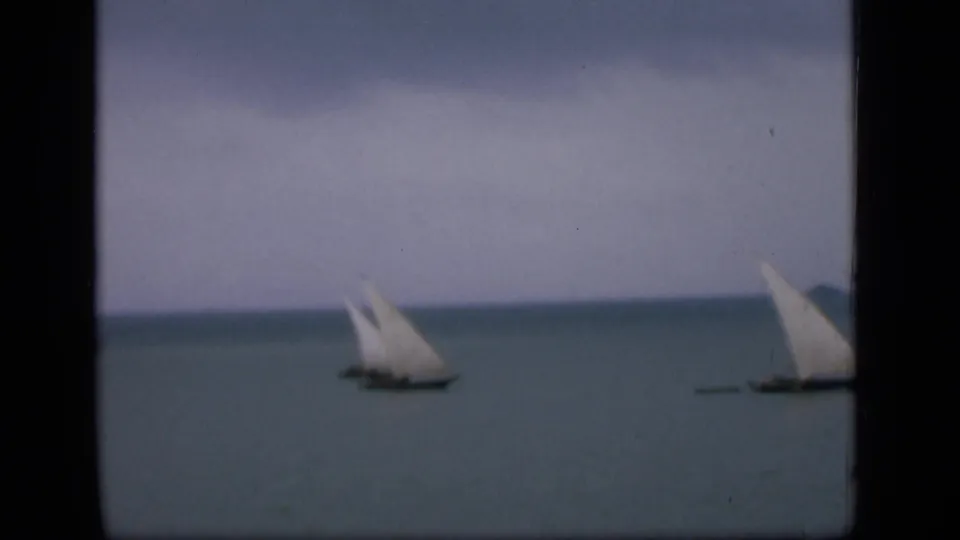Distant small sailboats with pale triangular sails on the water near Bombay, seen through soft 8mm framing.