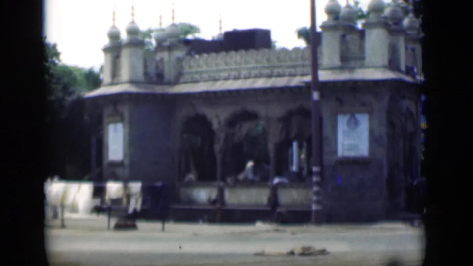 The camera holds on an ornate Bombay street building in 1946, showing arches, railings, pedestrians, and partial signage at frame edge.