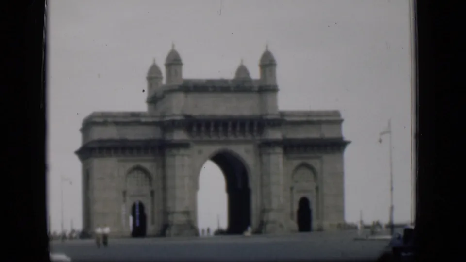 Home-movie frame of the Gateway of India in Bombay, showing the monument against a hazy sky with distant people on the pavement.