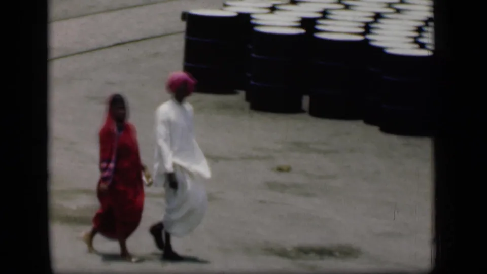 1946 Bombay 8mm frame showing a woman in red and a man in white passing stacked dark barrels.