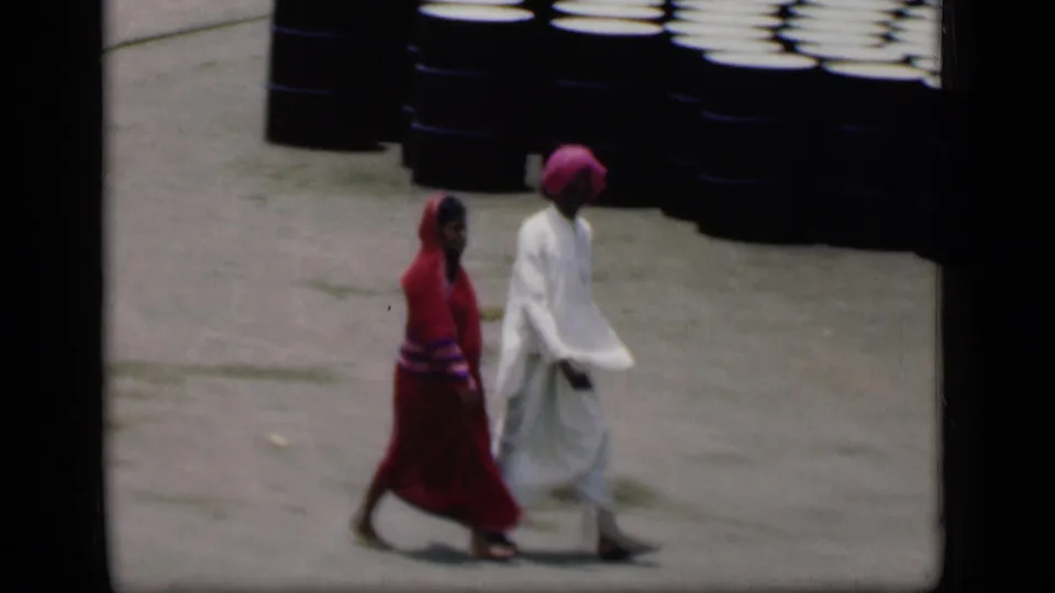 1946 Bombay frame of two pedestrians in traditional dress walking beside stacked dark barrels on pavement.