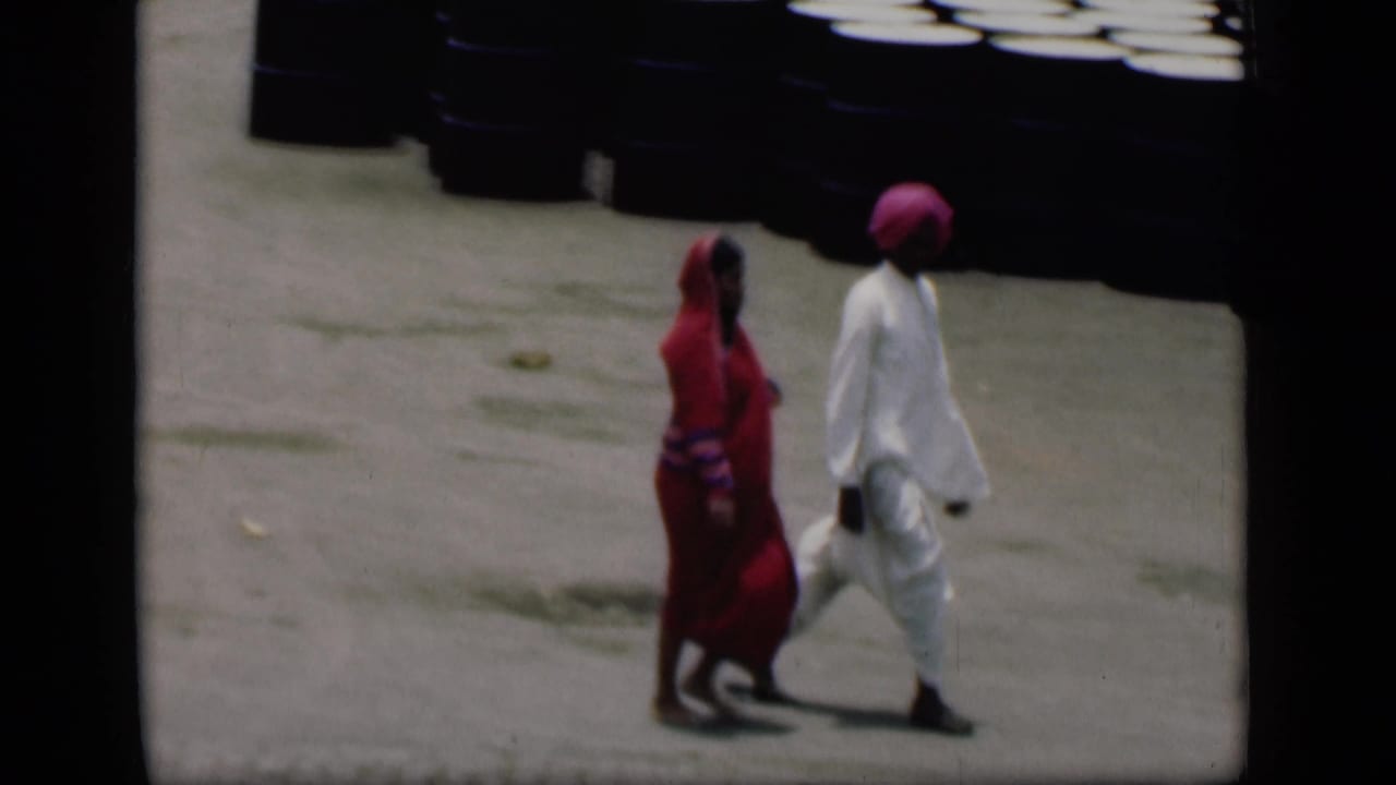 In 1946 Bombay home-movie footage, an adult in a red sari walks beside an adult in white clothing and a pink turban on pavement.