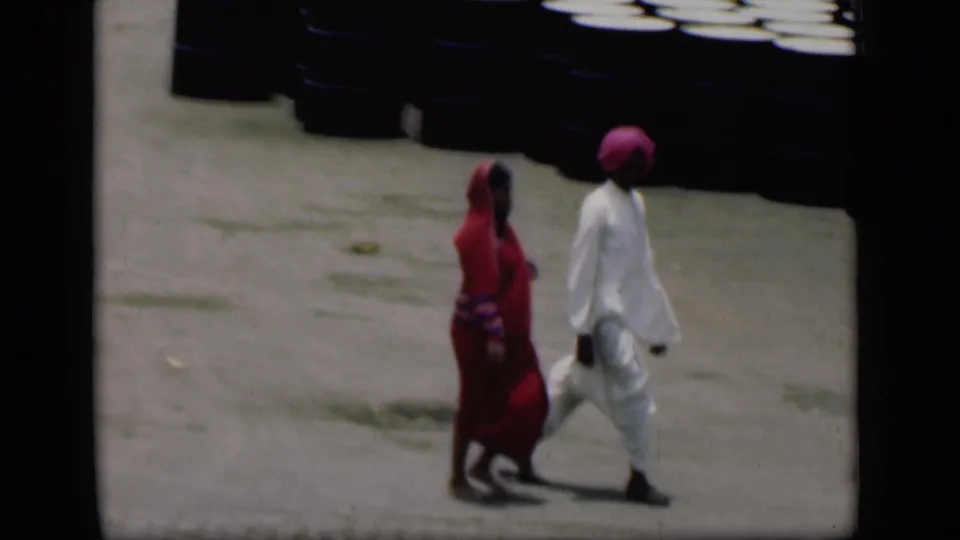 In 1946 Bombay home-movie footage, an adult in a red sari walks beside an adult in white clothing and a pink turban on pavement.