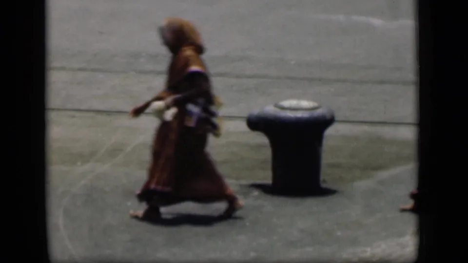 A person in red traditional clothing walks alone past a dark street bollard in the 1946 Bombay 8mm street scene.