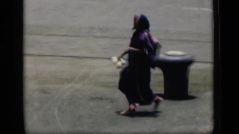 A pedestrian in dark traditional clothing crosses the frame near a low bollard in the 1946 Bombay home-movie street footage.