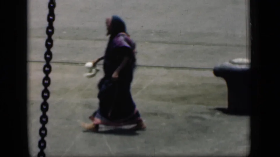 In 1946 Bombay home-movie footage, a woman in a dark sari and headscarf walks across a concrete street area.