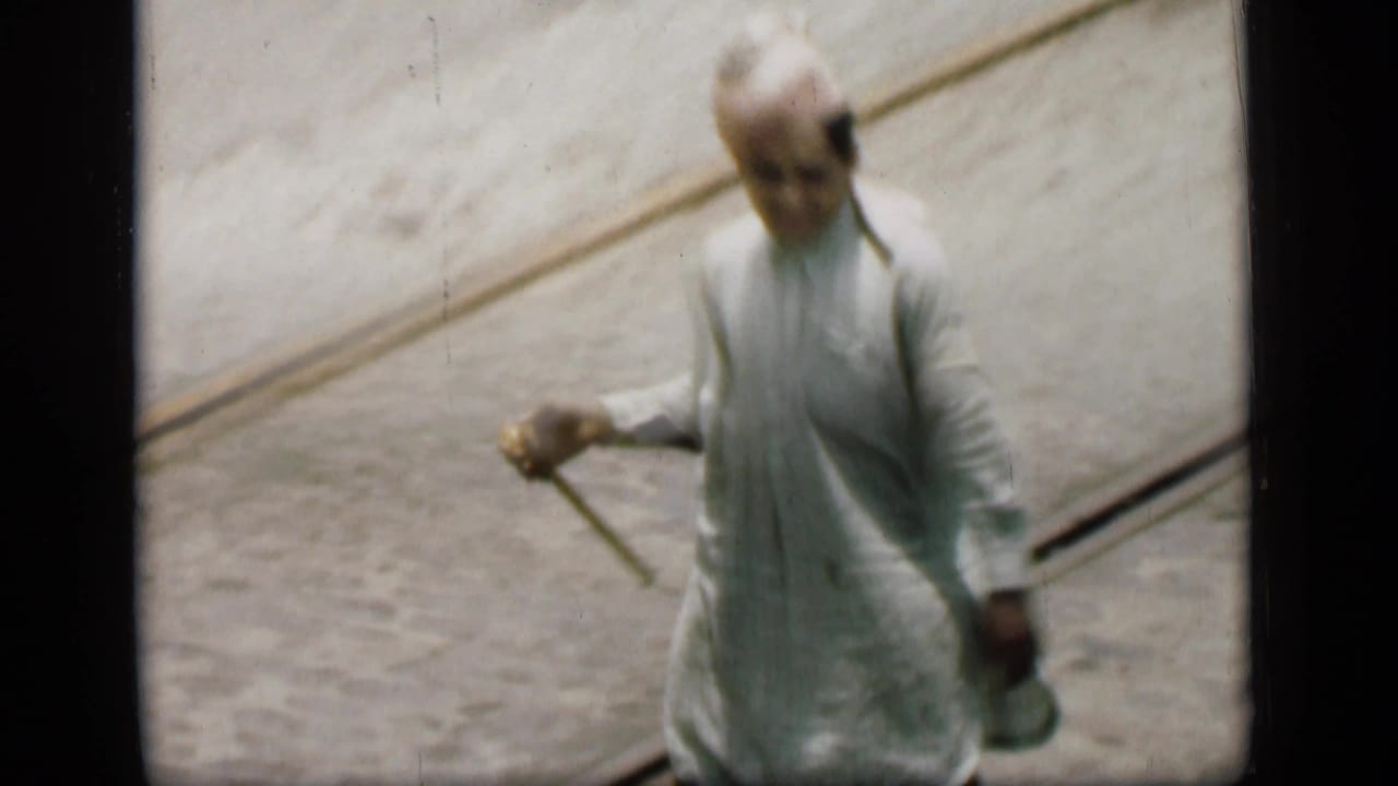 Man in pale clothing and turban walks with a cane beside tram tracks.
