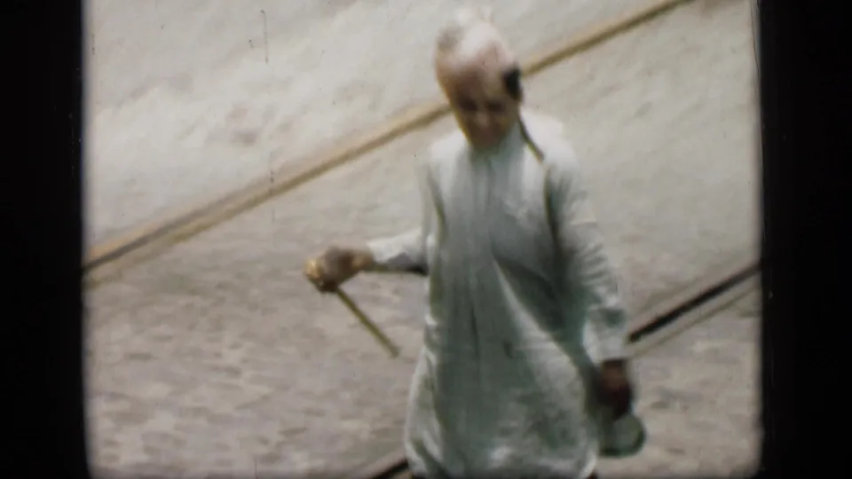 In 1946 Bombay, a man in a pale kurta and turban walks along a paved road with tram tracks, holding a cane.