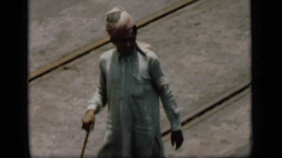 A man in a pale kurta and turban walks toward the camera on a Bombay street, with tram tracks visible behind him.