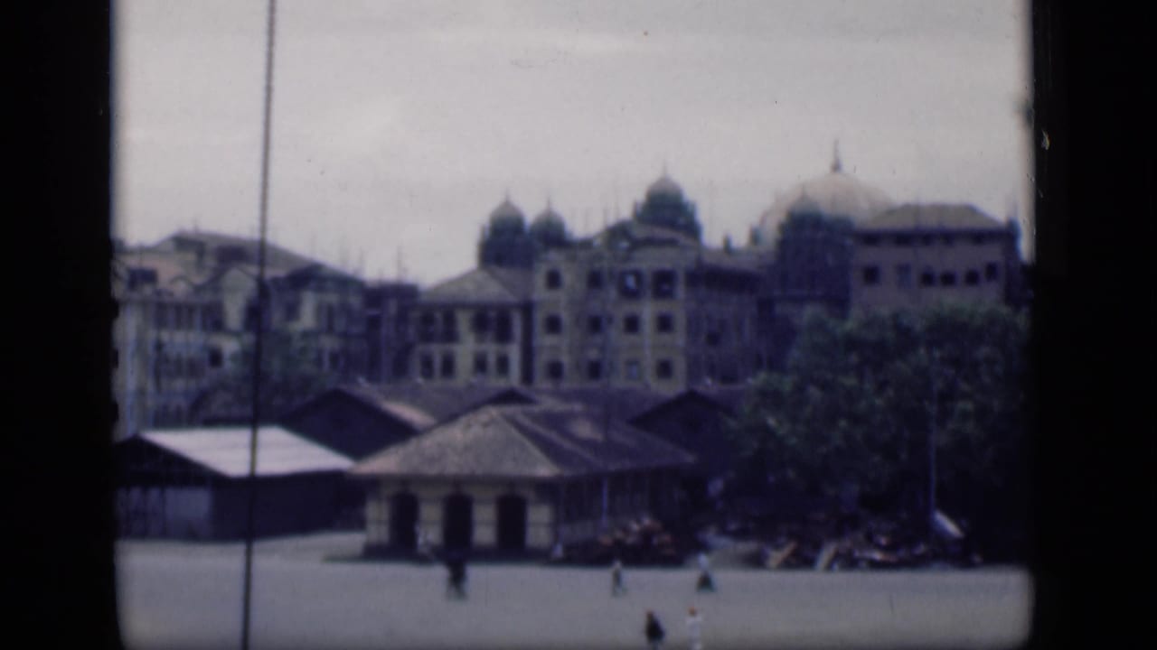 Wide 1946 Bombay plaza scene with domed architecture, foreground roofs, trees, and small walking figures.
