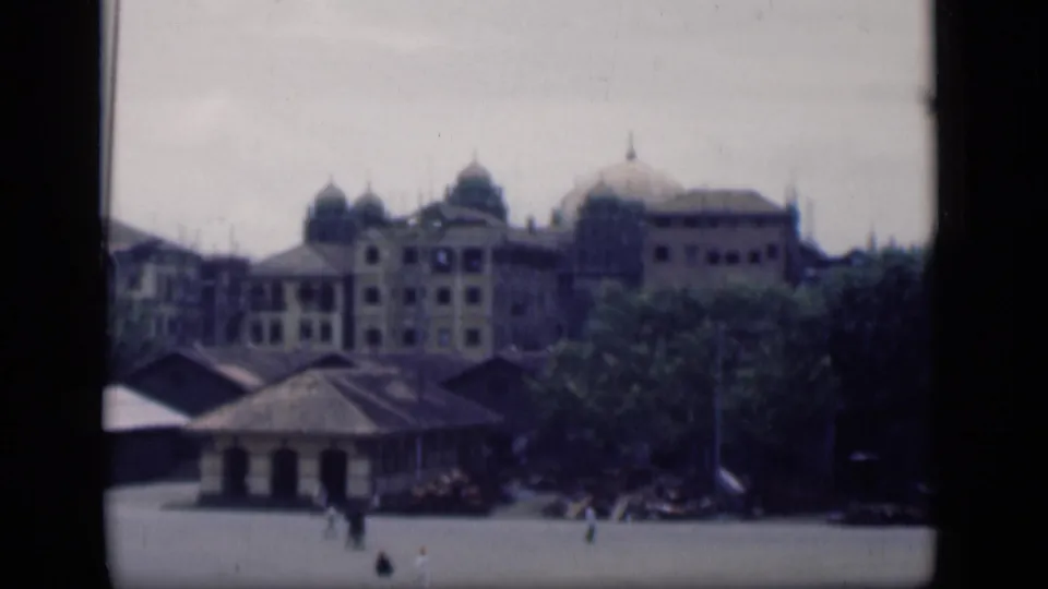 Wide 1946 Bombay exterior with domed colonial-era buildings, tree cover, and pedestrians scattered across the plaza.
